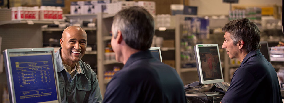 Two Ferguson associates at a plumbing supply counter assist a smiling plumbing contractor.