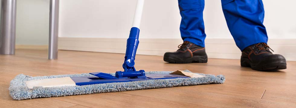 A facility worker wearing blue pants and brown work shoes dust mops a VCT floor.