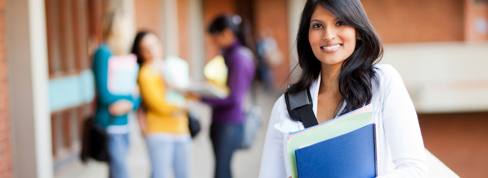 A student wearing a backpack and holding folders smiles at the camera; students talk in the background.