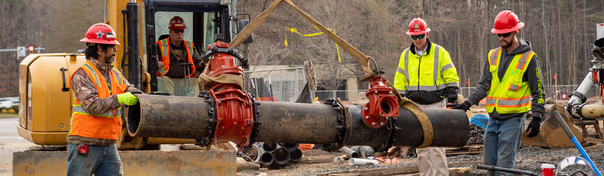 A waterworks utility contractor operates a front-end loader carrying an underground utility pipe, and three other contractors wearing reflective vests help guide it into place.